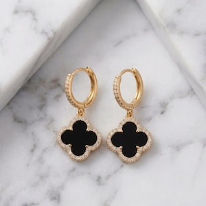 Peut inclure: Paire de boucles d'oreilles créoles dorées avec un pendentif en forme de trèfle à quatre feuilles. Le trèfle est noir avec une bordure de petits cristaux transparents. Présentées sur une surface en marbre blanc.