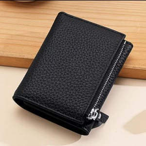 May include: Black leather wallet with a textured finish. It features a zip closure and a small tab. The wallet is closed and appears unused.