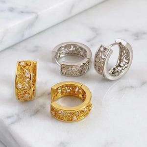 May include: Four hoop earrings, two gold and two silver, are presented on a marble surface. The gold earrings have an intricate filigree design with small, clear gemstones. The silver earrings also feature a filigree pattern.