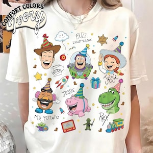 May include: Ivory-colored t-shirt featuring cartoon characters Woody, Buzz Lightyear, Jessie, Mr. Potato Head, Hamm, and Rex. The shirt has the words "Comfort Colors Ivory" in a circular design at the top left.