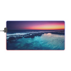 Op de afbeelding: Rechthoekige muismat met een zeelandschap van een zonsondergang boven de oceaan. De afbeelding toont tinten blauw, paars en oranje. De mat heeft een zwarte voedingsadapter aan de linkerkant en een oplichtende rand.