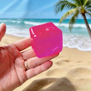 Op de afbeelding: Een helder roze, doorschijnend, geometrisch gevormd object wordt in een hand gehouden. De achtergrond toont een zandstrand, oceaangolven en een palmboom onder een blauwe lucht. Het object lijkt een decoratief item te zijn.