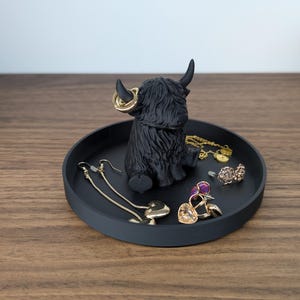 Op de afbeelding: Een zwart Highland koeienbeeldje met gouden oorringen staat in het midden van een ronde, matzwarte tray. De tray bevat diverse sieraden, waaronder oorbellen, ringen en een armband. Op de achtergrond staat een decoratieve pompoen.