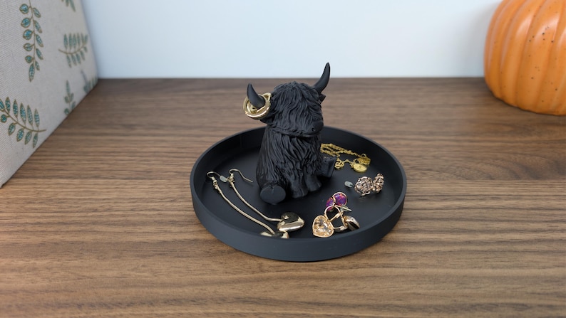 Op de afbeelding: Een zwart Highland koeienbeeldje met gouden oorringen staat in het midden van een ronde, matzwarte tray. De tray bevat diverse sieraden, waaronder oorbellen, ringen en een armband. Op de achtergrond staat een decoratieve pompoen.