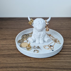Op de afbeelding: Een witte Highland-koeienfiguur met een ronde dienbladbasis, met gouden sieraden op zijn hoorns en dienblad. De sieraden omvatten kettingen, ringen en oorbellen. De dienblad staat op een houten oppervlak.