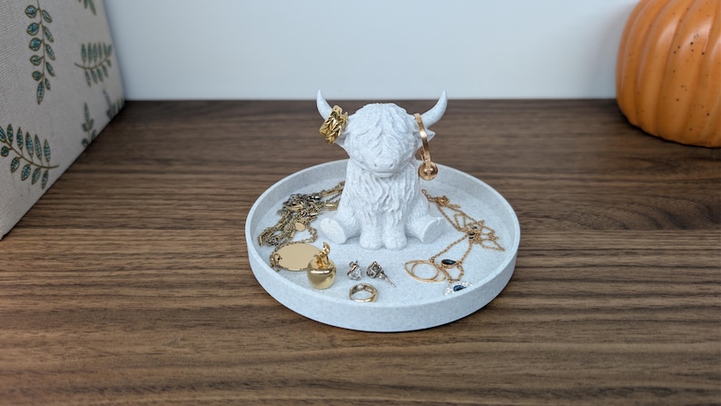 Op de afbeelding: Een witte Highland-koeienfiguur met een ronde dienbladbasis, met gouden sieraden op zijn hoorns en dienblad. De sieraden omvatten kettingen, ringen en oorbellen. De dienblad staat op een houten oppervlak.