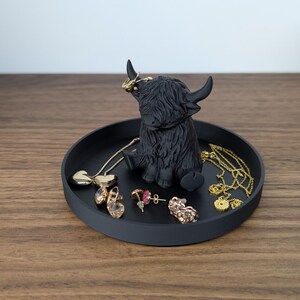 Op de afbeelding: Een zwarte ronde tray met een Highland koeienbeeldje in het midden, met diverse sieraden. De sieraden omvatten kettingen, oorbellen en ringen. De tray staat op een houten oppervlak.