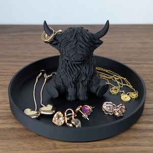 Op de afbeelding: Een zwarte Highland-koeienfiguur zit in een ronde, matzwarte tray. De tray bevat diverse goudkleurige sieraden, waaronder oorbellen en kettingen. De koe heeft gedetailleerde vacht en gebogen hoorns.