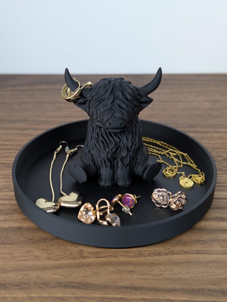 Op de afbeelding: Een zwarte Highland-koeienfiguur zit in een ronde, matzwarte tray. De tray bevat diverse goudkleurige sieraden, waaronder oorbellen en kettingen. De koe heeft gedetailleerde vacht en gebogen hoorns.