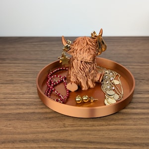 Op de afbeelding: Een koperkleurige metalen tray met een Highland koeienbeeldje in het midden. De tray bevat diverse sieraden, waaronder een rode kralenketting, gouden oorbellen en een gouden ketting. Op de achtergrond staat een oranje pompoen.