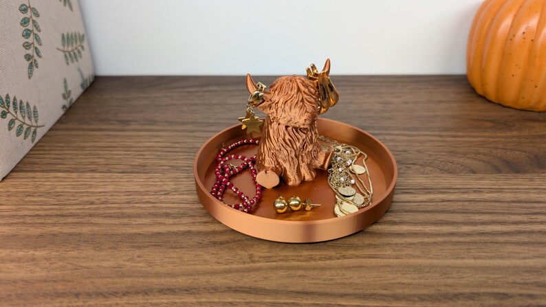 Op de afbeelding: Een koperkleurige metalen tray met een Highland koeienbeeldje in het midden. De tray bevat diverse sieraden, waaronder een rode kralenketting, gouden oorbellen en een gouden ketting. Op de achtergrond staat een oranje pompoen.