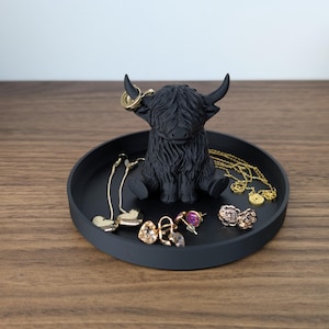 Op de afbeelding: Een zwart Highland-koeienbeeldje staat in het midden van een ronde, zwarte tray. De tray bevat diverse goudkleurige oorbellen en kettingen. Het beeldje heeft grote hoorns en een ruige vacht. Op de achtergrond is een pompoen te zien.