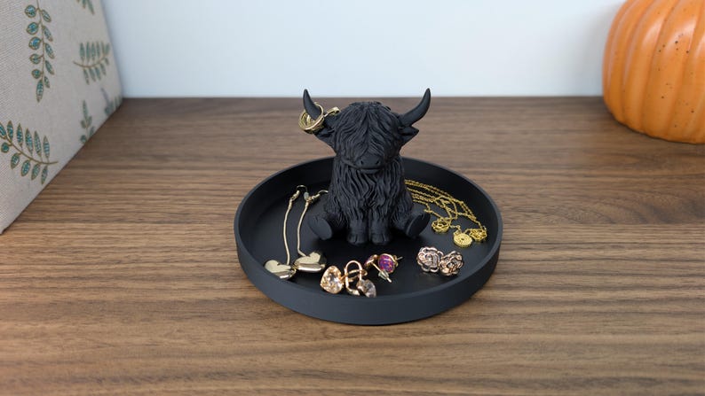 Op de afbeelding: Een zwart Highland-koeienbeeldje staat in het midden van een ronde, zwarte tray. De tray bevat diverse goudkleurige oorbellen en kettingen. Het beeldje heeft grote hoorns en een ruige vacht. Op de achtergrond is een pompoen te zien.