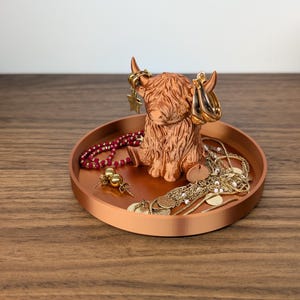 Op de afbeelding: Een koperkleurige tray met een Highland koeienbeeldje met oorbellen. De tray bevat diverse sieraden, waaronder kettingen, oorbellen en een kralenarmband. Het beeldje en de tray staan op een houten oppervlak.