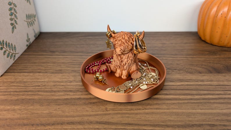 Op de afbeelding: Een koperkleurige tray met een Highland koeienbeeldje met oorbellen. De tray bevat diverse sieraden, waaronder kettingen, oorbellen en een kralenarmband. Het beeldje en de tray staan op een houten oppervlak.