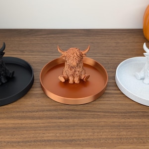 Op de afbeelding: Drie decoratieve trays, elk met een Highland koeienbeeldje. De trays zijn zwart, koper en wit. De koeienbeeldjes zijn zwart, koper en wit. Op de achtergrond staat een pompoen.