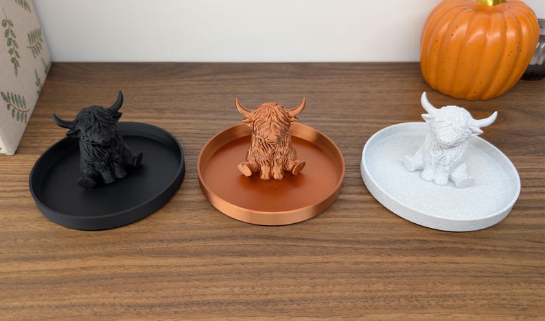 Op de afbeelding: Drie decoratieve trays, elk met een Highland koeienbeeldje. De trays zijn zwart, koper en wit. De koeienbeeldjes zijn zwart, koper en wit. Op de achtergrond staat een pompoen.