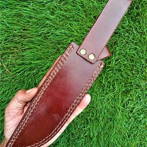 May include: A brown leather knife sheath with a strap and two brass-colored rivets. The sheath is hand-stitched with a contrasting white thread. The background is green grass.