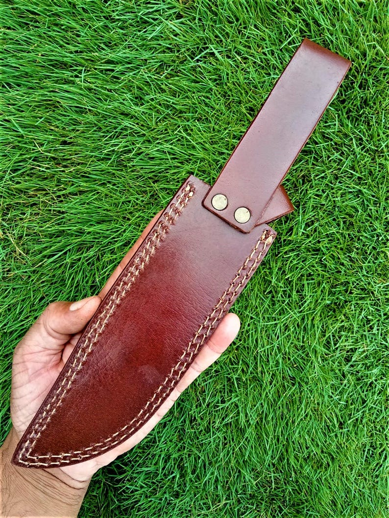 May include: A brown leather knife sheath with a strap and two brass-colored rivets. The sheath is hand-stitched with a contrasting white thread. The background is green grass.