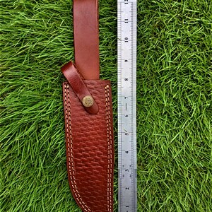 May include: A brown leather knife sheath with a woven texture and white stitching. The sheath has a belt loop and a snap closure with a decorative button. A ruler shows the sheath is approximately 12 inches long.