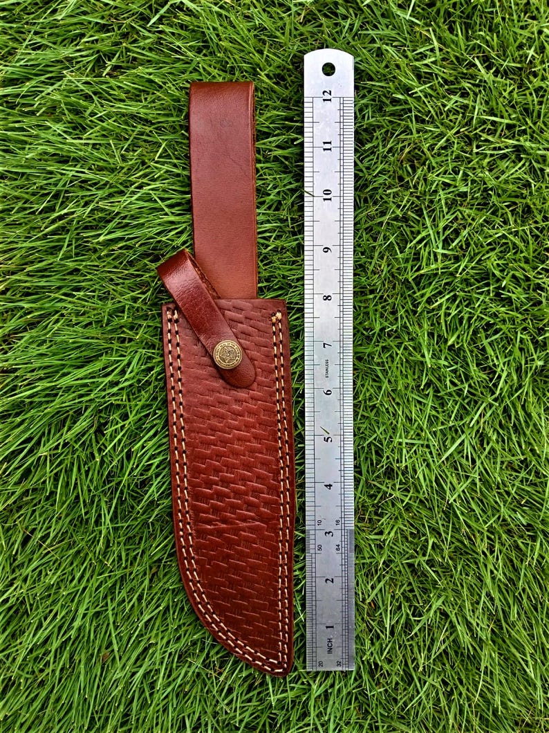 May include: A brown leather knife sheath with a woven texture and white stitching. The sheath has a belt loop and a snap closure with a decorative button. A ruler shows the sheath is approximately 12 inches long.