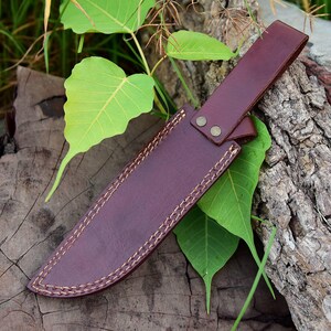 May include: A burgundy leather knife sheath with a strap and two brass rivets. The sheath is stitched with a contrasting light-colored thread. The knife sheath is resting on a wooden surface with green leaves.