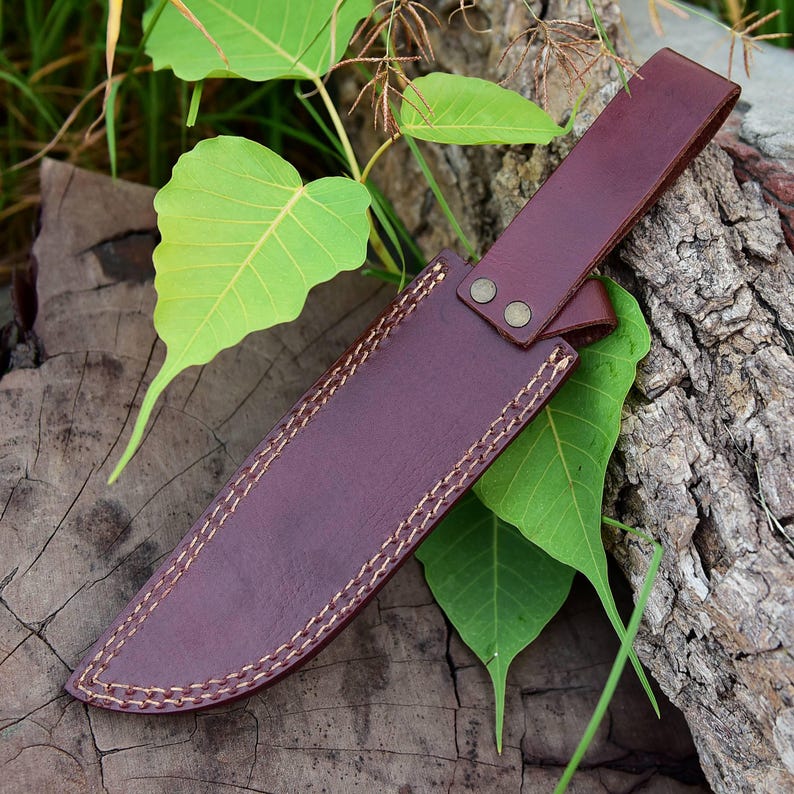May include: A burgundy leather knife sheath with a strap and two brass rivets. The sheath is stitched with a contrasting light-colored thread. The knife sheath is resting on a wooden surface with green leaves.