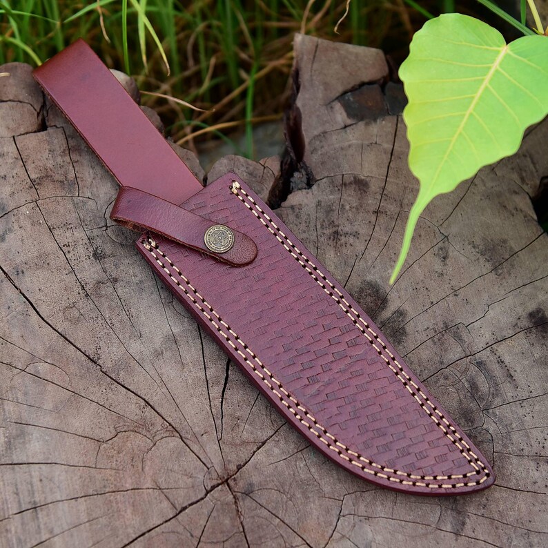 May include: A burgundy leather knife sheath with a woven texture and white stitching. The sheath has a strap with a button closure and is designed to hold a knife. The sheath is displayed on a wooden surface.