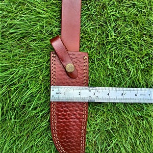 May include: A brown leather knife sheath with a textured basketweave pattern and white stitching. The sheath has a belt loop and a snap closure. The ruler shows the sheath is approximately 6 inches long.