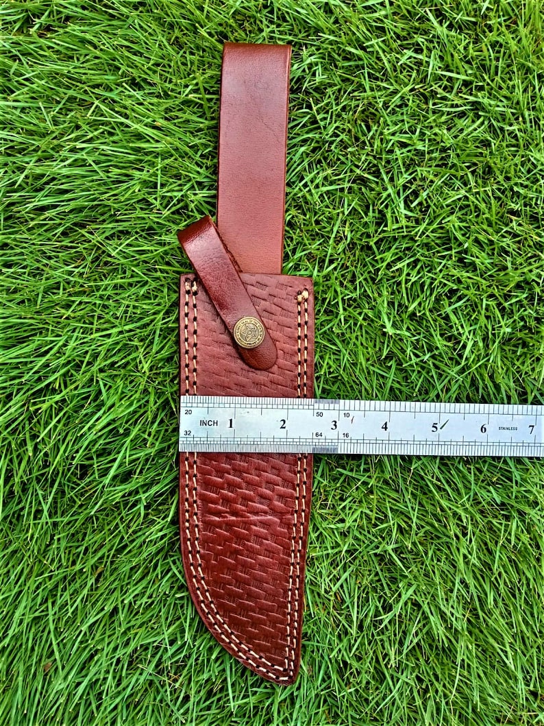 May include: A brown leather knife sheath with a textured basketweave pattern and white stitching. The sheath has a belt loop and a snap closure. The ruler shows the sheath is approximately 6 inches long.