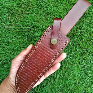 May include: A brown leather knife sheath with a textured pattern and white stitching. The sheath has a strap with a button closure. The background is green grass.