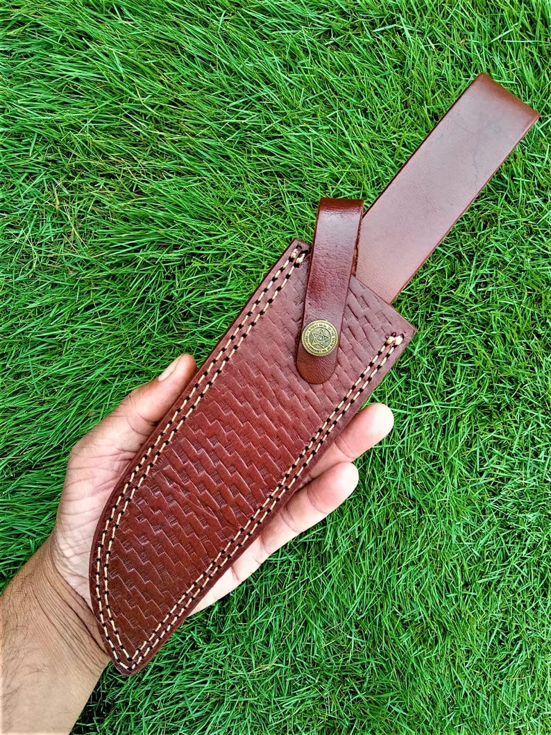 May include: A brown leather knife sheath with a textured pattern and white stitching. The sheath has a strap with a button closure. The background is green grass.
