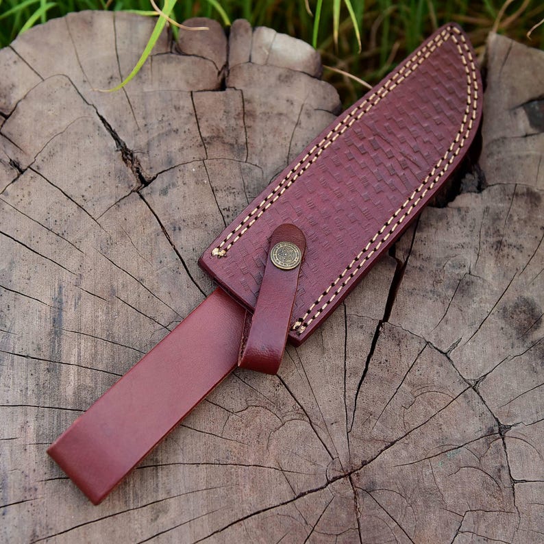May include: A handcrafted leather knife sheath in a rich burgundy color. The sheath features a woven texture and is secured with a leather strap and a decorative metal button. The edges are stitched with a contrasting light-colored thread.