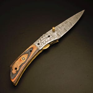 May include: A folding knife with a patterned steel blade and a wooden handle. The handle features a combination of brown, orange, and black wood, accented with brass hardware. The blade has a decorative etched design.
