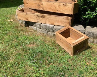 Op de afbeelding: Drie rechthoekige houten plantenbakken gestapeld op een stenen muur, met een vierde plantenbak in de buurt. De plantenbakken zijn gemaakt van verweerd hout, met natuurlijke nerven en knopen. De scène speelt zich buiten af op een grasveld.