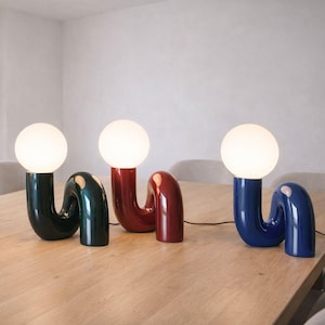 May include: Four modern table lamps with a curved design and spherical bulbs. The lamps are in white, dark green, red, and blue. They are placed on a light brown wooden table in a well-lit room.