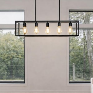 Op de afbeelding: Een moderne woonkamer met een zwarte rechthoekige lamp met vijf zichtbare gloeilampen. De kamer heeft twee grijze banken, een ronde salontafel en grote ramen met uitzicht op een groen landschap. De vloer is lichtgekleurd hout.