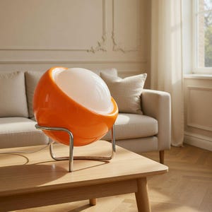 Peut inclure: Lampe de table orange de style rétro avec un abat-jour sphérique et une base en métal chromé. La lampe a un intérieur blanc et lumineux et repose sur une table en bois. C'est un luminaire décoratif.