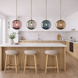 May include: Four pendant lights with spherical, ribbed glass shades in pink, gray, blue, and amber, each 20 cm in diameter, hang above a kitchen island. The kitchen features a white countertop, wooden cabinets, and three bar stools.