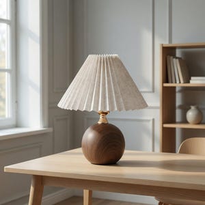 May include: A decorative table lamp with a pleated off-white lampshade and a round, dark brown wooden base. The lamp features a gold-colored neck and sits on a light-colored wooden table. The background includes a window, a bookshelf, and a chair.