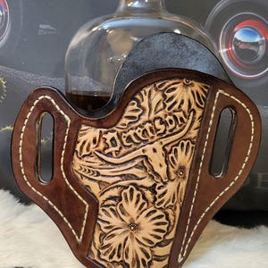 Può includere: Una fondina in pelle marrone fatta a mano con disegni floreali e longhorn, cucita con filo bianco. La fondina è ambientata su uno sfondo con una bottiglia di liquore e un'auto d'epoca.