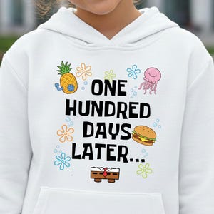 May include: White hooded sweatshirt with the text "ONE HUNDRED DAYS LATER..." and cartoon graphics of a pineapple, jellyfish, burger, and flowers. The sweatshirt has a front pocket.