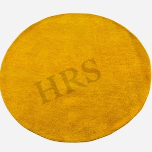 May include: A round, mustard-yellow rug with the letters "HRS" subtly imprinted in the center. The rug has a soft, textured appearance and is set against a plain white background. The rug is a home decor item.