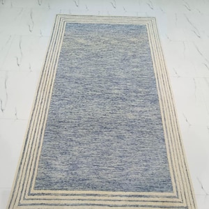 May include: Rectangular blue rug with a cream-colored border. The rug has a textured appearance, with a lighter blue center and a darker blue border. The cream border is made up of multiple parallel lines.