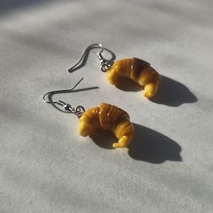 Puede incluir: Un par de pendientes colgantes con dijes de croissant en miniatura. Cada dije es de color marrón dorado con una textura realista de masa de hojaldre. Los croissants están unidos a ganchos de metal plateado para orejas perforadas.