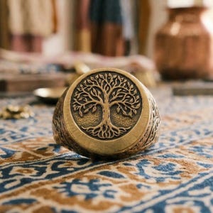 May include: A bronze ring featuring a tree of life design. The ring has a circular face with a detailed tree motif and an engraved band. The tree design includes a detailed trunk, branches, and leaves. The ring is set on a patterned surface.