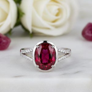 May include: A silver ring with a large, oval-cut ruby gemstone. The ruby is surrounded by a halo of smaller diamonds, and the band features a split shank with additional diamond accents. The ring is set against a blurred background of white flowers.
