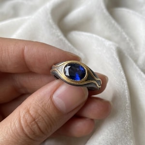 Op de afbeelding: Een zilveren ring met een gouden band en een grote, ovale, diepblauwe edelsteen. De ring heeft een gestructureerd, golvend ontwerp rond de edelsteen setting.