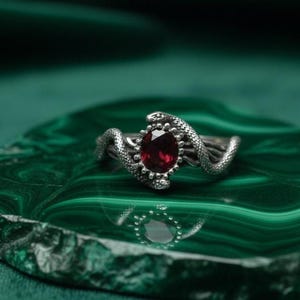 Puede incluir: Anillo de plata con una gema roja ovalada, flanqueada por dos diseños detallados de serpientes. El anillo se presenta sobre un fondo verde con patrones, creando un contraste llamativo.