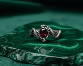 May include: A silver ring featuring a red oval gemstone, flanked by two detailed snake designs. The ring is set against a green, patterned background, creating a striking contrast.